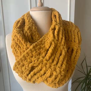 May include: A mustard yellow infinity scarf made of a soft, fuzzy yarn. The scarf is crocheted with a chunky stitch and has a loose, drapey texture.