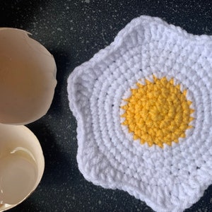 Puede incluir: Un posavasos de crochet blanco con forma de huevo frito con un centro amarillo. El posavasos está sobre una superficie negra junto a dos cáscaras de huevo rotas.
