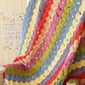 May include: A colourful crocheted blanket with a rainbow pattern. The blanket is made with a variety of different coloured yarns, including red, orange, yellow, green, blue, and purple.
