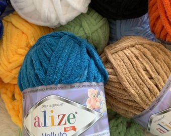 May include: A close-up of a variety of yarn balls in different colors, including teal, beige, green, white, orange, and yellow. The teal yarn ball has a label that reads "alize Velluto new" and "SOFT & BRIGHT".