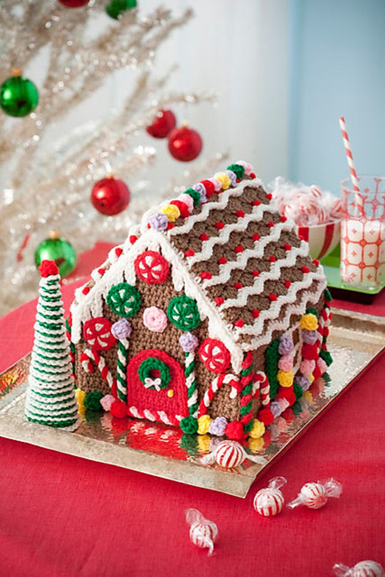 May include: A crocheted gingerbread house decorated with candy canes, gumdrops, and sprinkles. The house is sitting on a silver tray and surrounded by candy.