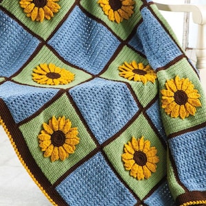 Puede incluir: Una manta de punto de crochet con un diseño de patchwork que presenta cuadrados de color azul, verde y marrón. Cada cuadrado tiene un girasol de crochet en el centro.