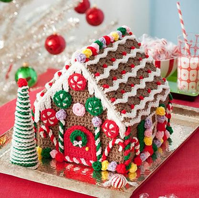 May include: A crocheted gingerbread house decorated with candy canes, gumdrops, and other festive details. The house is brown with white trim and a red door. It is sitting on a silver tray.