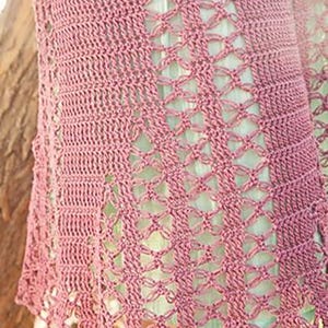 Puede incluir: Primer plano de una falda de ganchillo rosa polvoriento con un diseño geométrico calado. La falda tiene un dobladillo festoneado y una abertura lateral. El fondo es de color verde claro.