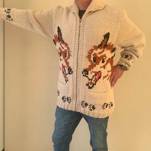 Puede incluir: Un cárdigan de punto color crema, de manga larga y con cremallera, que presenta un gráfico de lobo en tonos marrón, negro y rojo en los paneles frontales. El suéter incluye dos bolsillos delanteros, un cuello acanalado y huellas negras.