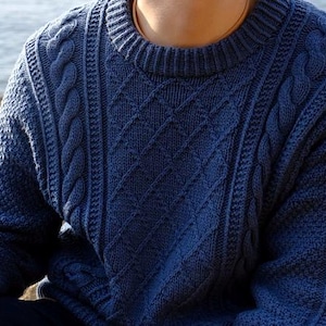 Könnte beinhalten: Ein marineblauer Rundhals-Pullover mit Zopfmuster. Der Pullover hat ein Rautenmuster auf der Vorderseite und den Ärmeln, mit Zopfmustern. Das Material ist weich und strukturiert. Ein klassischer Stil.