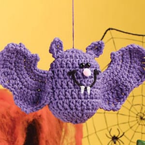 May include: Purple crocheted bat hanging ornament with black eyes and a pink nose. The bat has a toothy grin and is hanging from a string.