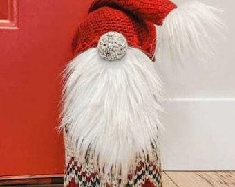 May include: A red and white crocheted gnome doorstop with a white fur beard and a red crocheted hat. The gnome is standing on a wooden floor.