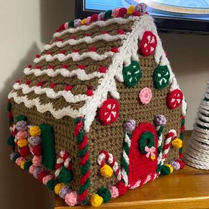 May include: A crocheted gingerbread house with white frosting, red candy canes, and colorful sprinkles. The house has a green door and a red roof.