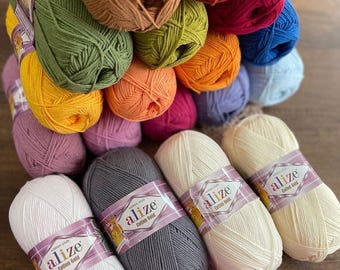 May include: A collection of yarn balls in various colors, including white, cream, gray, pink, orange, yellow, green, blue, red, and brown. The yarn balls are arranged in a pyramid shape and have the brand name "alize Cotton Gold" printed on the labels.