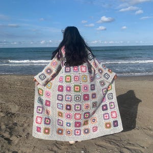 May include: A white crochet cardigan with a colorful granny square pattern. The cardigan is long and has a loose fit. The person wearing the cardigan is standing on a beach.