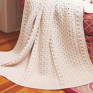 May include: A white crocheted throw blanket with a textured pattern. The blanket is draped over a chair with a red patterned fabric.