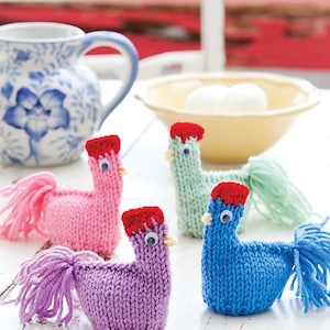 May include: Four knitted chicks in pink, purple, blue, and green with red combs and googly eyes. The chicks are sitting on a white table.