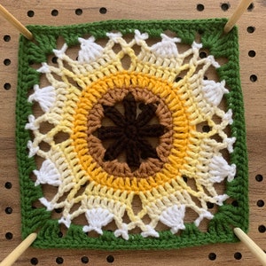 以下が含まれることがあります： 緑色の縁取りのある、かぎ針編みで作ったひまわりの正方形。ひまわりの中心は茶色で、花びらは黄色、白、クリーム色です。