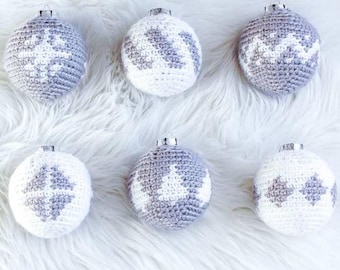 May include: Six handmade crochet Christmas ornaments. The ornaments are spherical, with a silver cap and a loop for hanging. Three are white with a gray snowflake design, and three are gray with a white snowflake design. They are arranged on a white, fluffy surface.