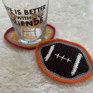 May include: Two crocheted coasters, one shaped like a football with brown, white, gray, and orange yarn, and the other a round coaster with yellow, orange, and red yarn. The text "Life is better with friends" is visible on a glass behind the coasters.