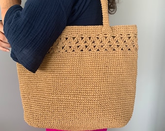 May include: A tan crocheted tote bag with a floral design on the top edge. The bag is made of a woven material and has a wide, flat base.