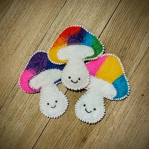 May include: Three felt mushroom ornaments with smiling faces. The mushrooms are in shades of purple, yellow, and rainbow.