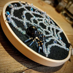 May include: A black spider with gold accents sits on a black felt background with a white beaded web design. The spider is in a round wooden frame.