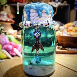 May include: A teal glass bottle with a white rope and blue glitter around the top. The bottle has a mermaid tail charm with a blue iridescent scale design hanging from it. The bottle is filled with white sand and a blue heart charm.
