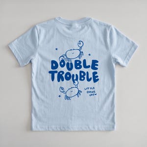 Puede incluir: Una camiseta de manga corta de color azul claro con un estampado gráfico en azul oscuro de dos cangrejos y el texto "DOUBLE TROUBLE" y "LITTLE CHAOS CREW" con pequeños detalles de estrellas. El diseño está centrado en la parte frontal.