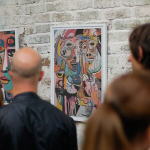 Puede incluir: Personas mirando pinturas de arte abstracto con formas geom&eacute;tricas y caras coloridas. Las pinturas est&aacute;n enmarcadas en blanco y colgadas en una pared de ladrillo blanco.