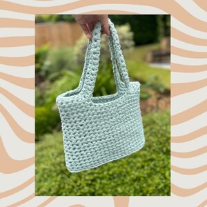 May include: A light green, hand-crocheted tote bag with two handles. The bag has a rectangular shape and a textured appearance. The background features green foliage and a decorative beige and white wavy pattern.