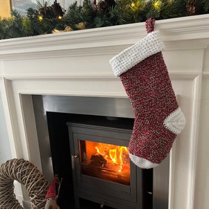 May include: A festive Christmas stocking, hand-crocheted with red, green, and white yarn. The cuff and toe are white. It hangs from a white mantelpiece adorned with pine and fairy lights. A fireplace with a burning fire is in the background.
