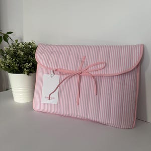 Könnte beinhalten: Eine rosa-weiß gestreifte Clutch mit passendem rosa Schleifenverschluss. Die Clutch hat eine abgerundete Klappe und ein Etikett mit der Aufschrift "Lot Lou". Der Artikel besteht aus einem weichen, gesteppten Material.