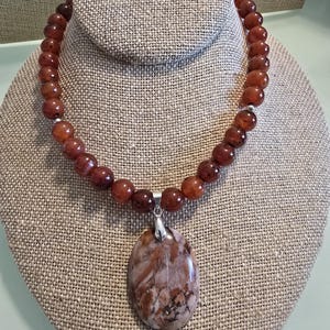 May include: A brown carnelian bead necklace with a large, oval-shaped pendant made of brown and white agate. The pendant is attached to the necklace with a silver bail.