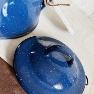 Puede incluir: Una cafetera y tapa de esmalte azul con motas blancas. La tapa tiene un asa negra. La olla está sujeta a una etiqueta con hilo. La olla está sobre una superficie blanca.