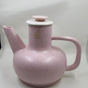 Puede incluir: Una tetera de cerámica rosa con un acabado moteado. La tetera tiene un cuerpo redondeado, un asa curva y un pico. La tapa es blanca con un pomo rosa estriado. La tetera está diseñada para servir té u otras bebidas calientes.