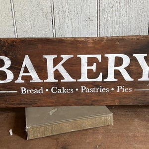 May include: A distressed wooden sign, featuring the word "BAKERY" in white capital letters. Beneath, the sign lists "Bread • Cakes • Pastries • Pies." The sign is a dark brown colour, suggesting a vintage or rustic aesthetic.