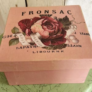 May include: A square, light pink box featuring a large, dark red rose illustration on the lid. The lid includes black printed text reading FRONSAC, ROSA SINENSIS, I:APAYRO, LIBOURNE, and other partial words, along with a small crown graphic.