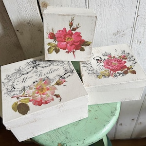 May include: Three white wooden boxes with floral designs. The boxes feature pink and red roses with green leaves. One box has the text "Paris". The boxes are stacked on a green stool.