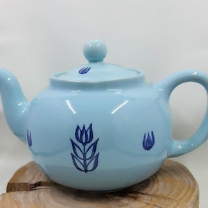 Pode incluir: Uma chaleira azul claro com uma tampa branca e desenhos de tulipas azuis. A chaleira tem um corpo redondo e um bico lateral.