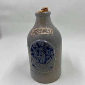 May include: A small, gray ceramic bottle with a cork stopper. The bottle has a blue and white label that reads "Zeer Oude Genever" and a blue and white image of a windmill and a man in traditional Dutch clothing. The bottle is marked "Gebr. De Windt A.D. 1690".