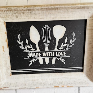 Könnte beinhalten: Ein gerahmtes Tafelschild mit einem weißen Holzrahmen im Used-Look. Die schwarze Tafel zeigt weiße Illustrationen eines Löffels, eines Schneebesens und eines Spatels mit dem Schriftzug "MADE WITH LOVE".
