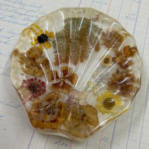 Puede incluir: Un plato decorativo de resina en forma de concha con flores secas incrustadas. Las flores son en tonos de amarillo, naranja, rojo y blanco. La resina es transparente, lo que permite ver las flores. El plato mide aproximadamente 10 cm de ancho.