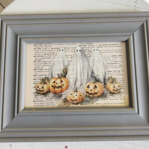 May include: A rectangular gray wooden frame containing a print of three white ghosts standing behind four orange jack-o'-lanterns. The artwork is layered over a background of dictionary text definitions starting with the word decide.