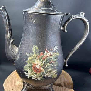 Könnte beinhalten: Eine silberne Teekanne mit einem Blumendesign mit einer roten Rose und grünen Blättern. Die Teekanne hat einen weißen Knopf auf dem Deckel und drei Füße.