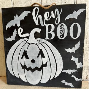 May include: A black wooden Halloween sign with white painted accents. The sign features a smiling pumpkin, bats, and the words "hey BOO". A jute rope is attached for hanging. The sign is a festive decoration for the Halloween season.