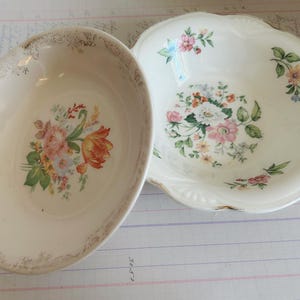 May include: Two vintage porcelain serving bowls. One is oval-shaped with a floral design featuring pink, orange, and white flowers. The other is round with a scalloped edge and a similar floral pattern. Both have gold trim.