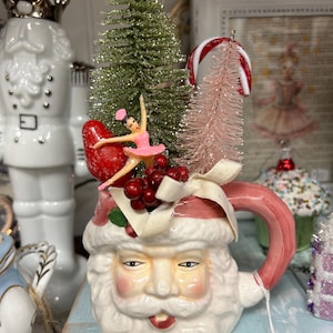 May include: A ceramic Santa Claus mug with a pink handle and a white beard. The mug is decorated with a small pink Christmas tree, a red bird, a candy cane, and a ballerina figurine.