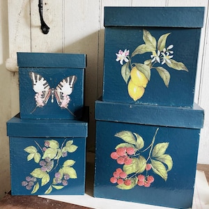 Pode incluir: Quatro caixas de armazenamento quadradas, empilhadas, azul-petróleo, com tampas. Cada caixa apresenta um design diferente: uma borboleta, limões, amoras e framboesas. As caixas estão sobre uma superfície de madeira. Medidas em centímetros.