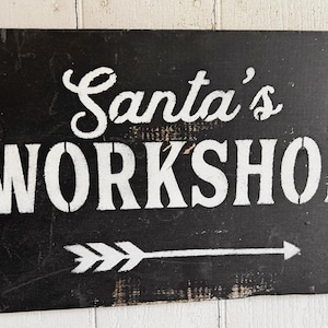 Peut inclure: Panneau en bois noir avec l'inscription blanche "Santa's WORKSHOP" et une flèche pointant vers la droite. Le panneau a un aspect vieilli, suggérant un style rustique ou vintage, et est probablement utilisé pour les décorations de Noël.