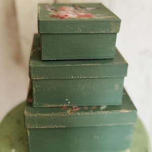 May include: Three stacked, rectangular boxes in varying sizes. The boxes are painted a muted green with distressed edges. The top box features a floral design with pink and red roses. The boxes are arranged on a green surface.