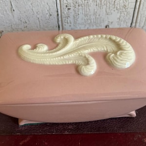 May include: A rectangular, pink ceramic jewelry box with a cream-colored, raised, decorative swirl design on the lid. The box has a curved base and small feet.