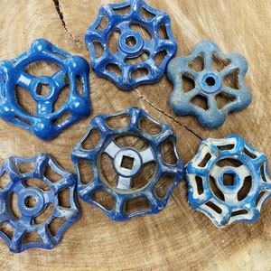 May include: Six vintage blue water valve handles with a starburst design. The handles are made of metal and show signs of wear, with some paint chipping. The handles are arranged on a light brown wooden surface.