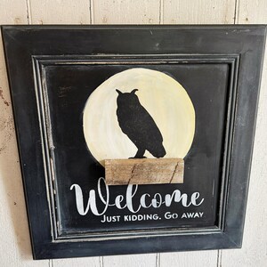 Könnte beinhalten: Ein schwarz gerahmtes Schild mit schwarzem Hintergrund, das eine Silhouette einer Eule zeigt, die auf einem Holzabsatz vor einem gelben Mond sitzt. Auf dem Schild steht "Welcome" über "Just Kidding. Go Away" in weißer Schrift, ein humorvolles Wohnaccessoire.